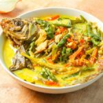 Semangkuk kari ikan mas dengan kuah santan kental berwarna kuning keemasan, dilengkapi potongan tomat dan kentang