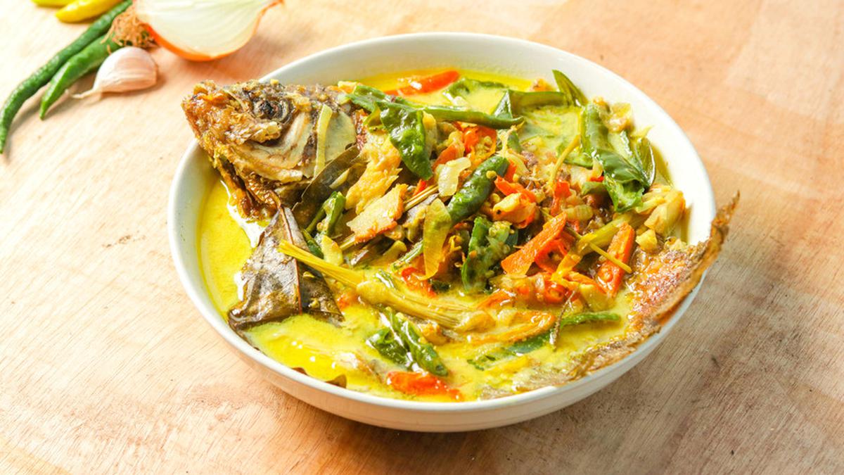 Semangkuk kari ikan mas dengan kuah santan kental berwarna kuning keemasan, dilengkapi potongan tomat dan kentang